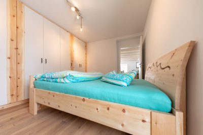 Ein Schlafzimmer im Ostallgäu. Ein Kleiderschrank mit cremeweiß lackierten Türen mit Zirbenfries. Das Kopfteil vom Bett ist mit Panoramaausschnitt.